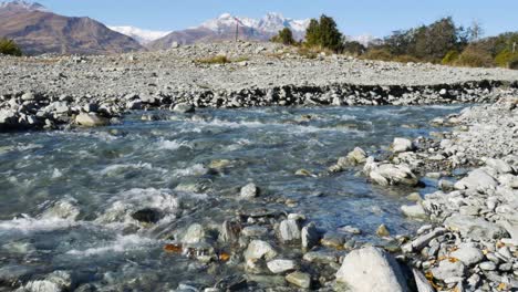 Primer-Plano-En-ángulo-Bajo-Del-Río-Que-Fluye-Entre-Piedras-Y-Rocas-Durante-La-Pista-De-Quema-De-Glaciares-En-Nueva-Zelanda---Montañas-Nevadas-En-El-Fondo