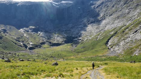 Toma-Aérea-De-Arriba-Hacia-Abajo-De-Un-Excursionista-Caminando-Cuesta-Abajo-Impresionante-Paisaje-Montañoso-En-Verano---Parque-Nacional-De-Fiordland,-Nueva-Zelanda