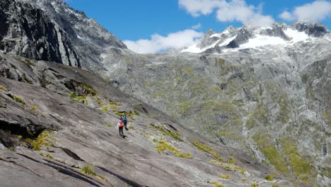 Grupo-De-Excursionistas-Caminando-En-La-Montaña-Rocosa-En-El-Camino-De-La-Silla-De-Montar-De-Gertrude-En-El-Parque-Nacional-De-Fiordland-Durante-El-Verano---Picos-Nevados-De-La-Montaña-En-El-Fondo