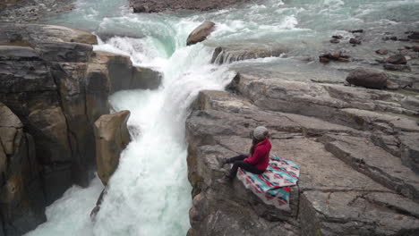Mujer-Sentada-En-Una-Roca-Sobre-Una-Cascada-Y-Un-Río-Glacial-En-El-Campo-Canadiense-En-Un-Frío-Día-De-Otoño