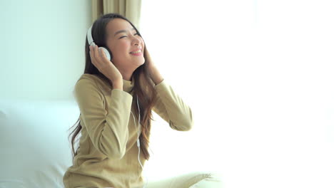 Happy-Energetic-Asian-Girl-Listening-to-Music-from-Overhead-Headphones-While-Sitting-on-Bed-near-Window-Smiling---slow-motion