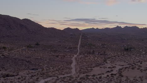 Aerial-of-desert-landscape-in-Joshua-Tree,-California-at-sunset-with-a-lonely-dirt-road-going-straight-back-and-into-the-hills