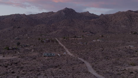 Flyover-homes-and-dirt-road-towards-hills-of-Joshua-Tree-National-Park-in-California-at-sunset-on-a-beautiful-evening