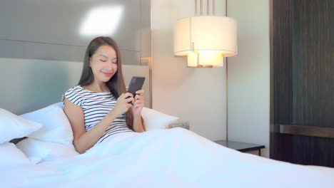 A-cute-young-woman,-sitting-up-in-a-big-comfy-bed-relaxing-as-she-texts-on-her-smartphone