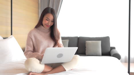 An-attractive-young-woman-sits-on-the-bed-of-her-hotel-suite-as-she-enters-data-into-her-laptop