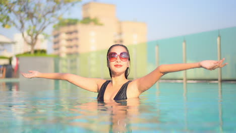 Eine-Schöne-Frau-In-Einem-Resort-Schwimmbad-Hebt-Ihre-Arme,-Um-Den-Morgen-Zu-Begrüßen