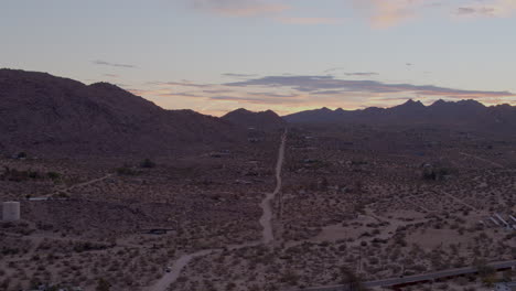 Aerial-push-towards-and-over-lonely-dirt-road-heading-towards-the-hills-of-Joshua-Tree-National-Park-in-California-on-a-beautiful-evening-at-sunset