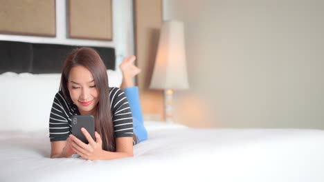 While-lying-on-her-stomach-on-the-bed,-a-pretty-young-woman-focuses-on-her-smartphone-and-smiles