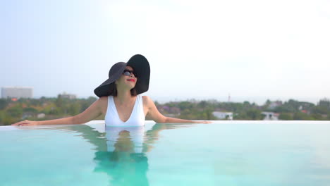 A-woman-in-a-huge-black-floppy-sun-hat-leans-along-the-edge-of-a-swimming-pool-as-she-takes-in-the-view