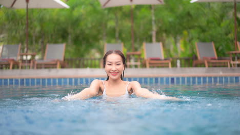 Woman-playfully-splashing-water-with-hands-towards-the-camera-inside-swimming-pool-at-Hotel-Lounge---slow-motion-front-view