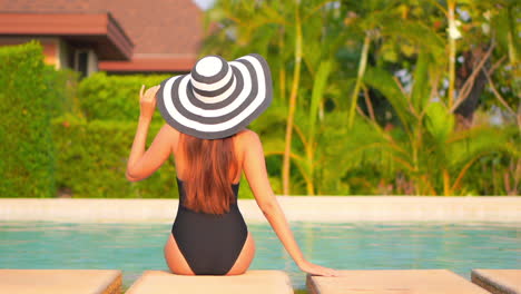 Woman-sitting-on-the-edge-of-the-swimming-pool-at-an-exotic-hotel-in-Florida-wearing-black-monokini-and-striped-hat-and-touching-hat-border,-back-view-slow-motion-handheld
