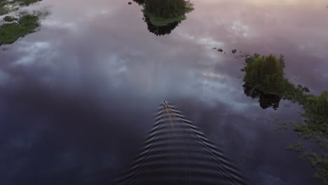 Aerial-Drone-shot-of-small-motorboat-with-2-passangers-cruising-slowly-in-a-calm-lake-surface-by-golden-hour-sunset