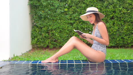 Profile-of-a-young,-fit-woman-in-a-one-piece-bathing-suit-sits-on-the-edge-of-a-swimming-pool-as-she-reads-a-book