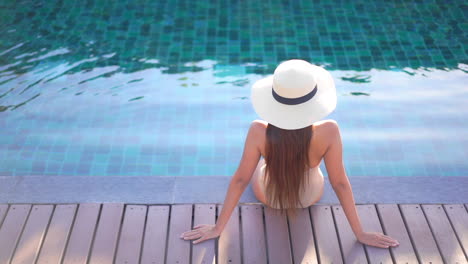 Fashionable-woman-in-sun-hat-relaxing-sitting-on-the-edge-of-swimming-pool---back-view