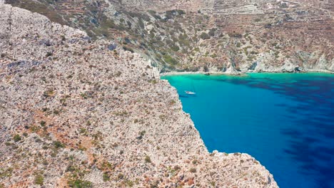 Aerial-Flying-through-Rock-cliff-reveal-Pristine-turquoise-Vorina-Beach,-Boat-Anchored
