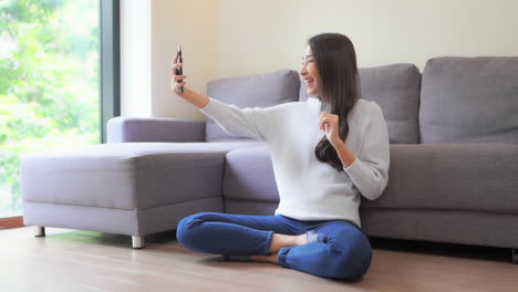 Glad-Asian-woman-with-a-pleasant-smile,-waves-with-a-hand-at-camera-of-cell-phone-in-home-interior,-makes-video-call-to-her-best-friend-sitting-on-a-floor