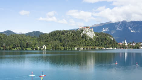 Antena---Gente-En-Paddleboards,-Lago-Bled,-Alpes-Julianos,-Eslovenia,-Pan-Ascendente-A-La-Izquierda