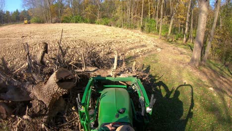 High-angle-point-of-view-on-small-green-tractor-using-lift-forks-to-pickup-a-tree-stump-from-a-pile-and-move-debris-near-woods-in-early-autumn