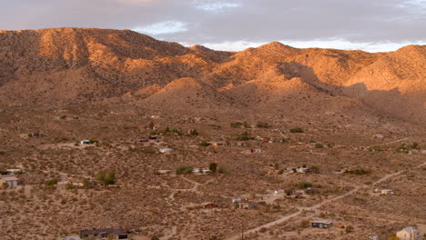 Aerial-view-of-little-houses-and-dirt-road-in-Joshua-Tree-at-sunrise-on-a-pretty-morning-with-jagged-hills-in-the-background