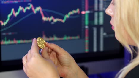 Blonde-woman-holding-and-looking-at-bitcoin-in-her-hands-in-front-of-cryptocurrency-trading-graphs