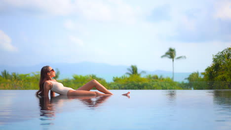 A-beautiful,-very-fit-woman-enjoys-the-sun-as-she-reclines-in-the-shallow-water-of-a-resort-infinity-pool