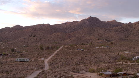 Flyover-a-dirt-road-in-the-dessert-over-houses-and-towards-jagged-hills-in-Joshua-Tree,-California-at-sunrise