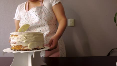 Latin-woman-wearing-an-apron-preparing-cooking-baking-a-cake-spreading-butter-frosting-with-a-white-cake-scraper