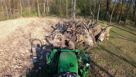 High-angle-point-of-view-on-small-green-tractor-using-lift-forks-to-pickup-a-tree-stump-from-a-pile-and-move-debris-near-woods-in-early-autumn