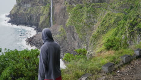 Hooded-man-walks-out-to-overlook-revealing-Véu-da-Noiva-waterfall,-slow-motion
