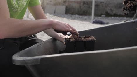man-puts-soil-from-wheelbarrow-to-plastic-container-with-bare-hands,-closeup