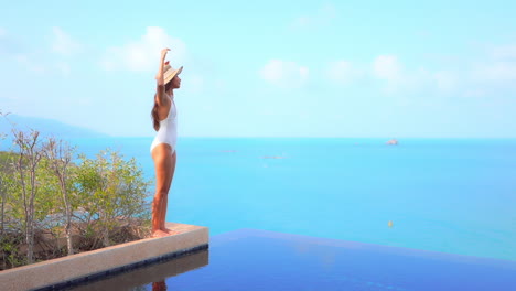 While-standing-on-the-edge-of-an-infinity-pool-looking-out-over-the-ocean-raises-her-arms-in-joy