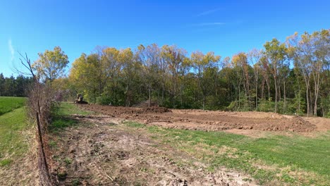 Wide-angle-view-of-a-yellow-bulldozer-leveling-soil-at-a-construction-site-with-wooded-area-in-background-in-early-autumn