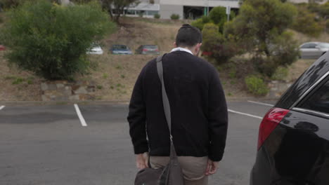A-male-office-worker-retrieves-his-bag-from-the-back-of-his-car-and-places-it-over-his-shoulder-as-he-walks-across-a-car-parking-lot-to-his-work-place-office