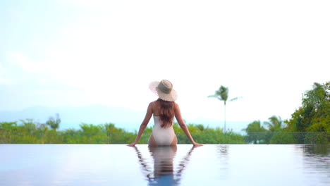 A-healthy,-fit,-sexy-woman-sits-on-the-edge-of-a-resort-infinity-pool-with-her-back-to-the-camera-as-she-looks-out-over-the-ocean