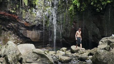 Young-fit-man-walking-over-rocks-away-from-small-lush-jungle-waterfall