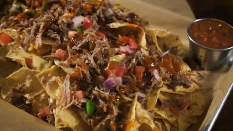 Nacho-pile-of-corn-tortilla-chips-slathered-with-queso-cheese-sauce-pulled-pork-and-pico-de-gallo-salsa,-close-up-slider-4K