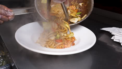 Chef-uses-tongs-to-plate-steaming-hot-freshly-stir-fried-udon-noodles-on-white-plate,-slow-motion-HD