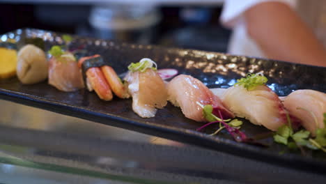 Eine-Bunte-Auswahl-An-Traditionellen-Nigiri-sushi-Auf-Einer-Schwarzen-Platte-In-Einem-Japanischen-Restaurant-Mit-Koch-Im-Hintergrund,-Schieber-Nahaufnahme-Hd