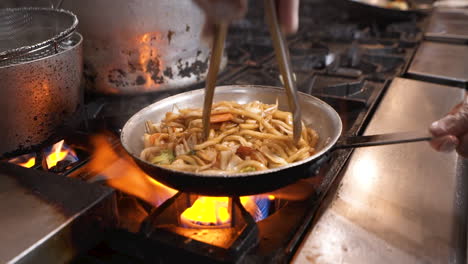 Chef-uses-tongs-to-flip-and-toss-traditional-udon-noodles-with-shrimp-and-vegetables-in-steel-pan-over-fiery-commercial-stove,-slow-motion-HD