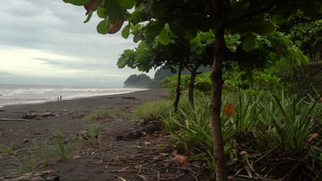 Ein-Paar-In-Der-Ferne-Spaziert-Am-Ufer-Eines-Schwarzen-Sandstrandes-In-Costa-Rica-Entlang---Verschiebbarer-Weitwinkelblick-Auf-Die-Atemberaubende-Landschaft