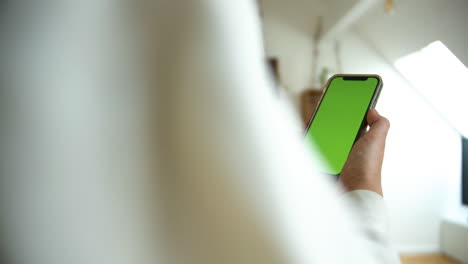 Side-view-of-a-causal-dressed-man-holding-smartphone-with-green-screen-chroma-key-in-white-home-background