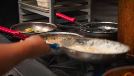 El-Chef-Lanza-Para-Incorporar-Pasta-Fresca-En-Salsa-De-Crema-Alfredo-Sobre-La-Estufa-De-La-Cocina-Del-Restaurante-Italiano,-Sartenes-Con-Diferentes-Salsas-Alineadas-En-La-Estufa-De-Preparación,-Cámara-Lenta-4k
