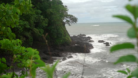 Amazing-view-of-a-rocky,-tropical-shoreline-with-ocean-waves-crashing-against-the-rocks-and-cliff---sliding-view-through-the-jungle-flora