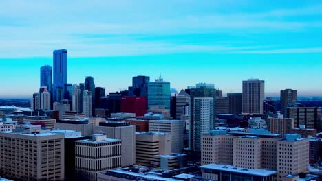 Winter-Snow-Covered-congested-skyscraper-buildings-post-modern-architecture-in-Downtown-Edmonton-where-smoke-stacks-are-blowing-in-a-freezing-cold-blue-hour-of-a-day-with-some-clouds-and-blue-horizon