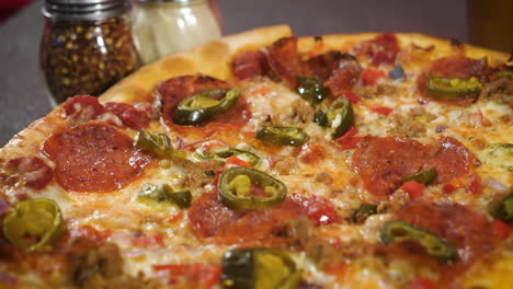 Thin-crust-whole-pepperoni-pizza-with-jalapenos,-grated-cheese-and-crushed-red-pepper-flakes-in-background,-slider-close-up-4K