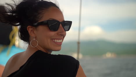 Beautiful-young-woman-wearing-sunglasses,-smiling,-and-enjoying-a-sailboat-trip-along-a-tropical-coast