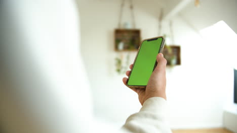 A-man-holding-a-smartphone-with-green-screen-in-vertical-in-white-home-background