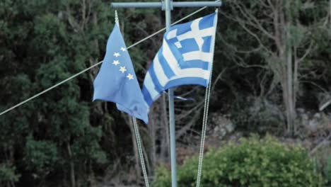 European-Union-And-Greek-Flag-Hanging-Side-By-Side-Waving-In-The-Wind-With-Background-Of-Trees-In-Kefalonia,-Greece