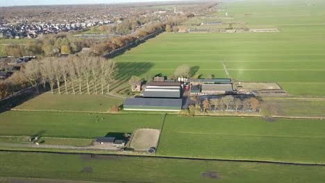 Antenne-Eines-Bauernhofs-Mit-Sonnenkollektoren-Auf-Dem-Dach-Einer-Scheune-Im-Ländlichen-Holland---Drohne-Fliegt-Rückwärts-Und-Enthüllt-Grüne-Wiesen