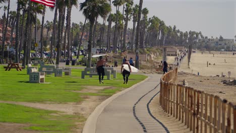 Surfistas-Que-Llevan-Tablas-De-Surf-A-Lo-Largo-Del-Carril-Bici-En-Huntington-Beach,-California